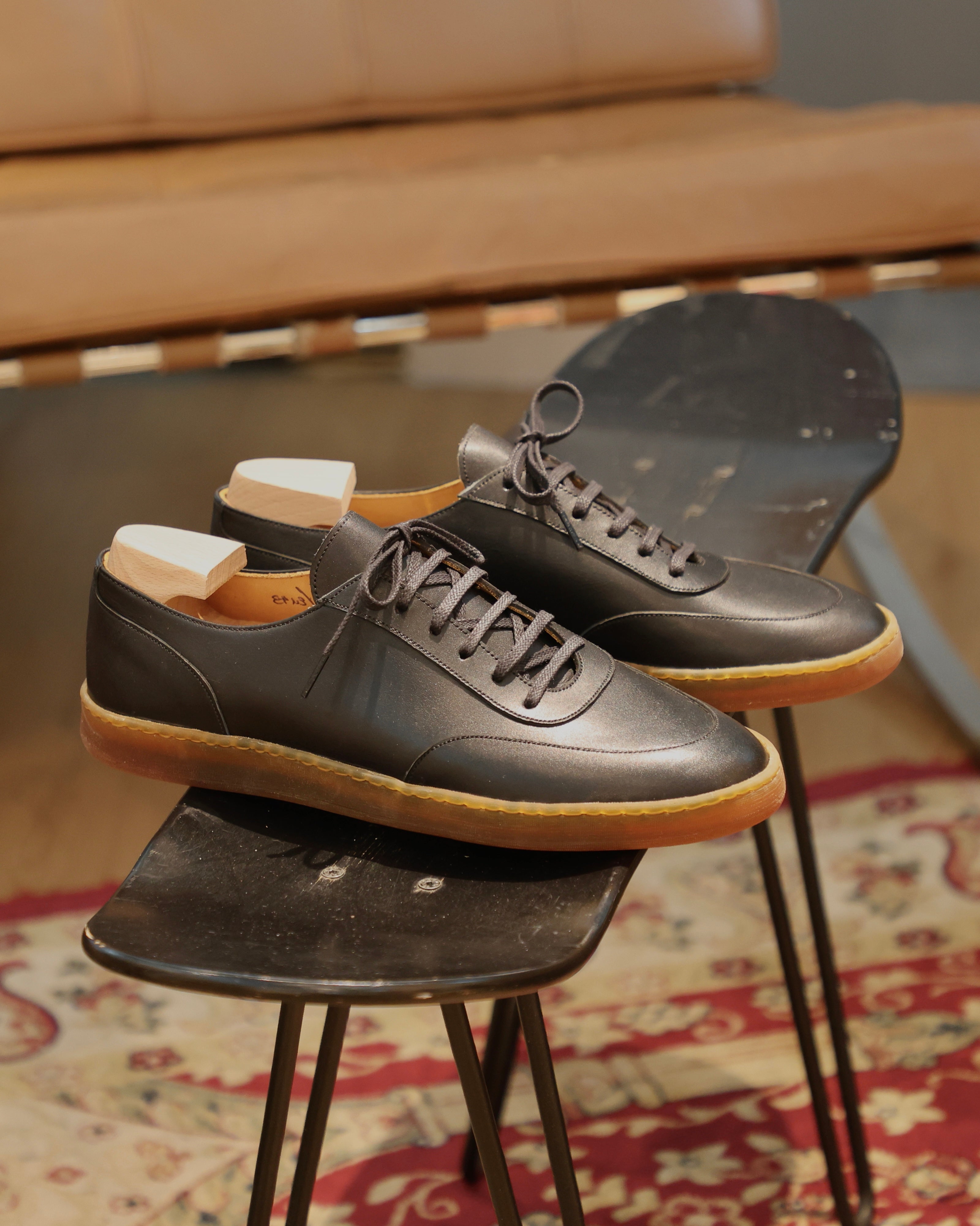 A pair of blackleather lace-up shoes with gum soles, displayed on black metal stools in an indoor setting with a patterned rug and leather furniture in the background.