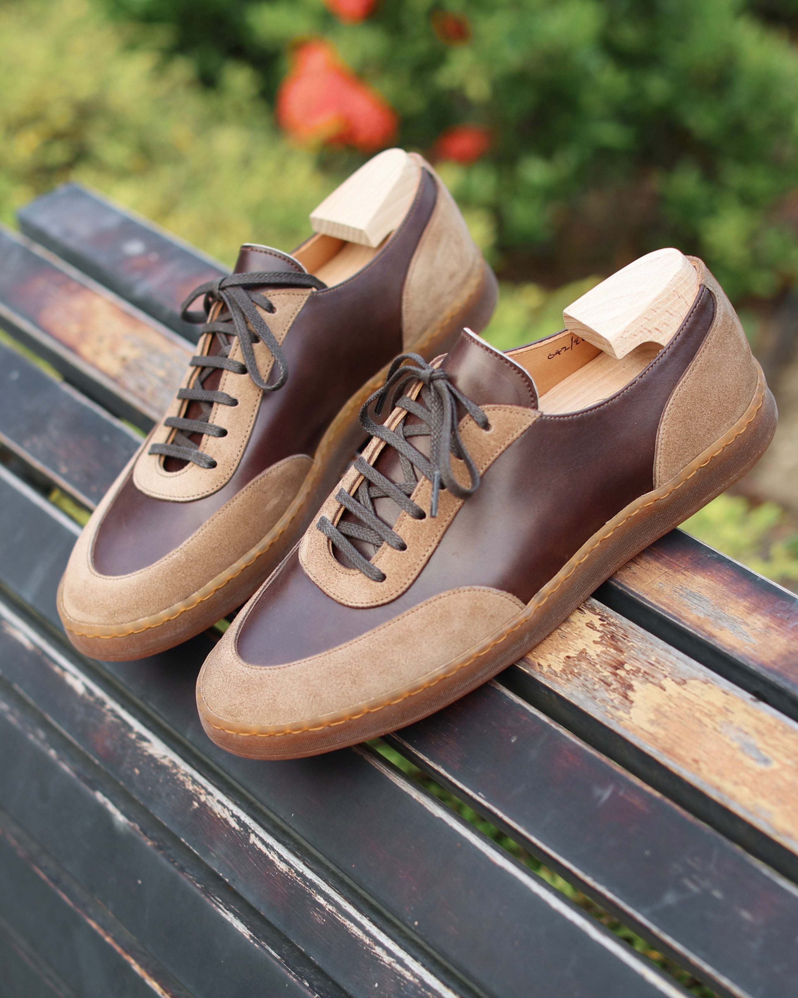 A pair of brown leather and suede lace-up shoes with light tan soles, displayed on a weathered wooden bench with a blurred green garden background.